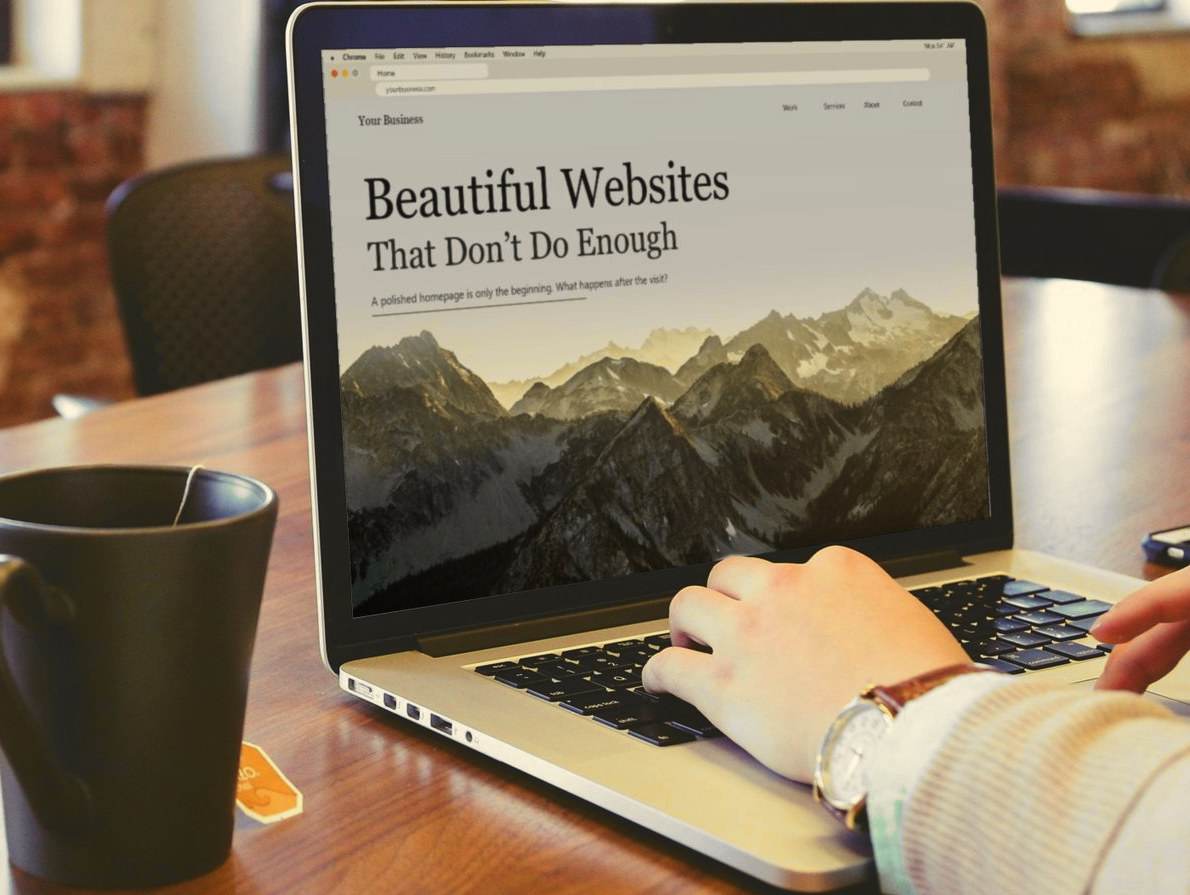 A laptop on a wooden desk, its screen showing a website that reads: Beautiful Websites, That Don’t Do Enough.